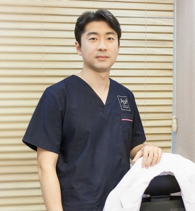 에이펙스 로고가 달린 남색 수술복을 입은 송현석 대표원장이 흰 가운이 걸린 의자 옆에 서서 정면을 보고 있습니다.