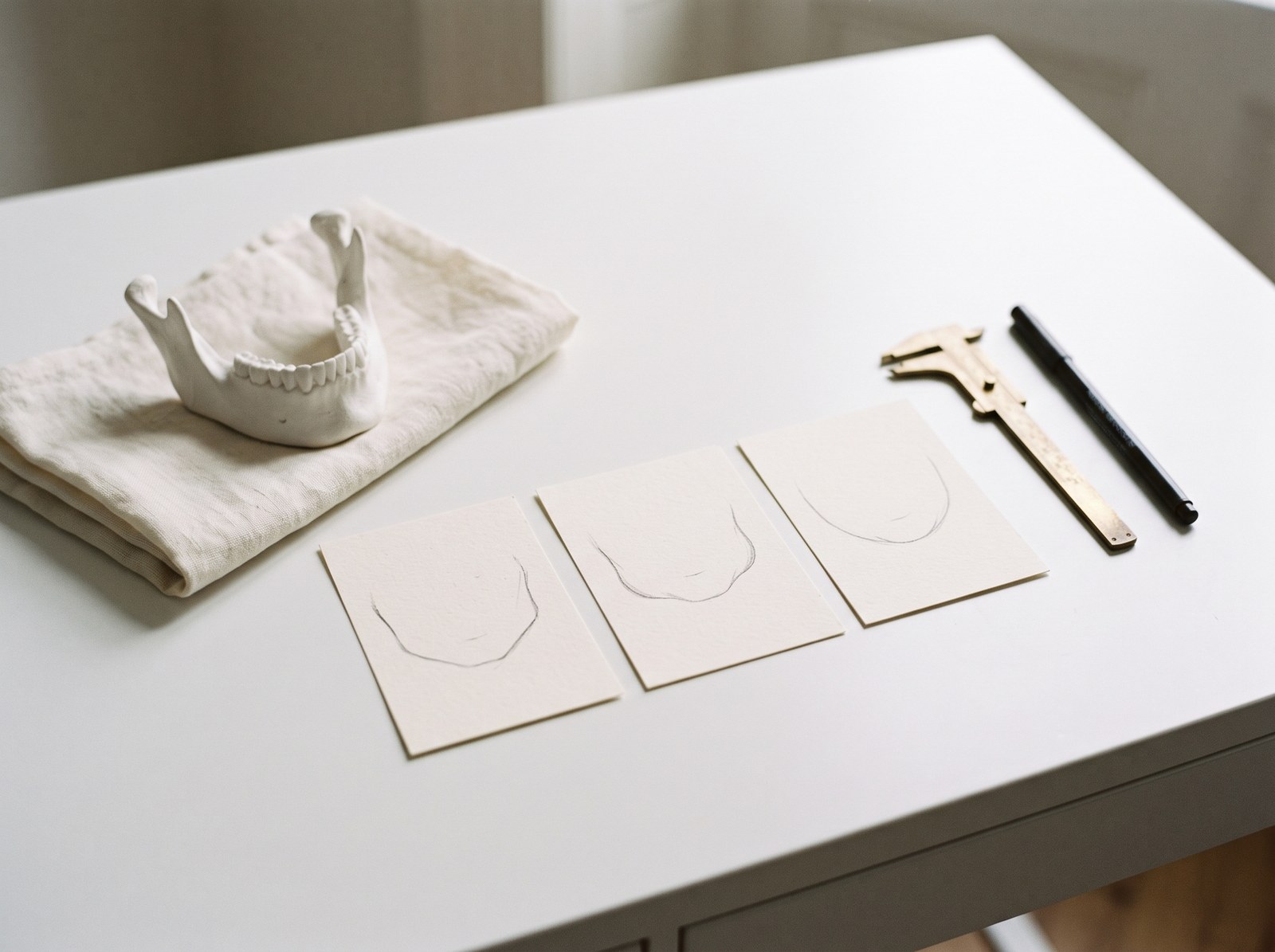 A white plaster model of a lower jawbone rests on a cream-colored cloth over a white desk, alongside three cards with pencil-sketched jawlines, a brass caliper, and a marker pen.