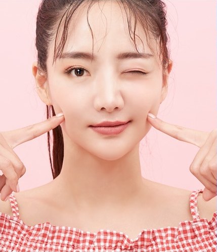 분홍색 배경에서 양 검지손가락으로 볼을 가리키며 윙크하는 여성의 얼굴 지방흡입 관련 클로즈업 모습