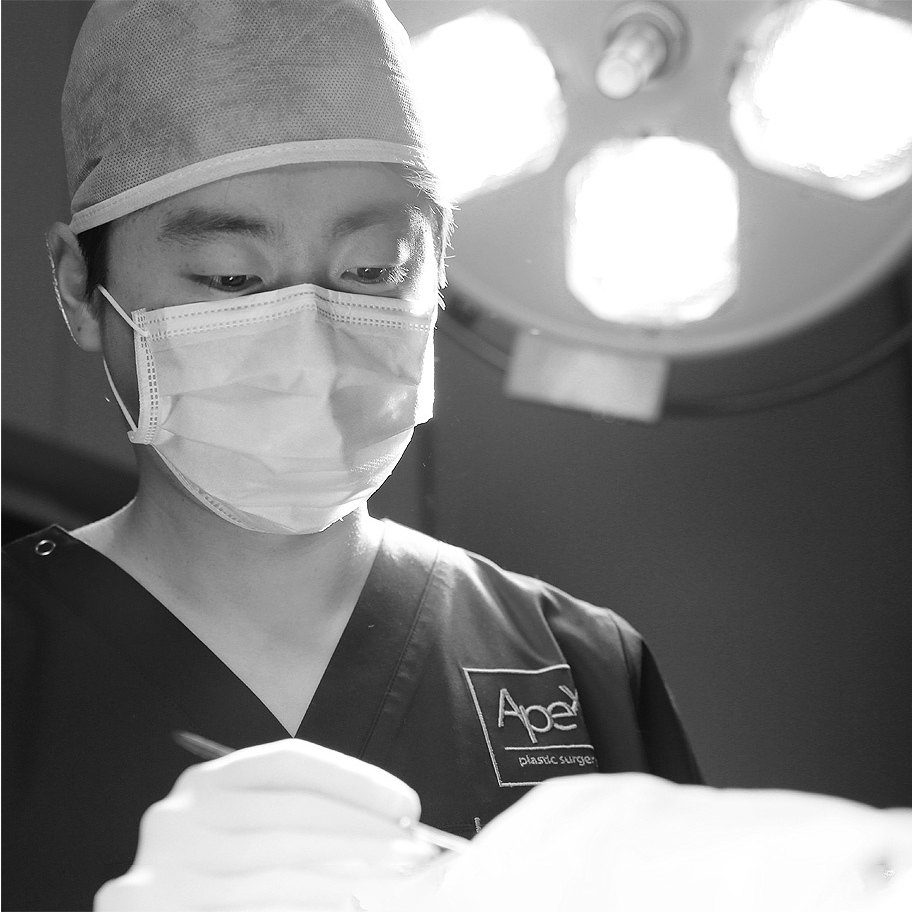 수술용 조명 아래에서 마스크와 수술모를 착용하고 긴코·화살코 성형 집도에 집중하는 에이펙스성형외과 의료진의 흑백 모습