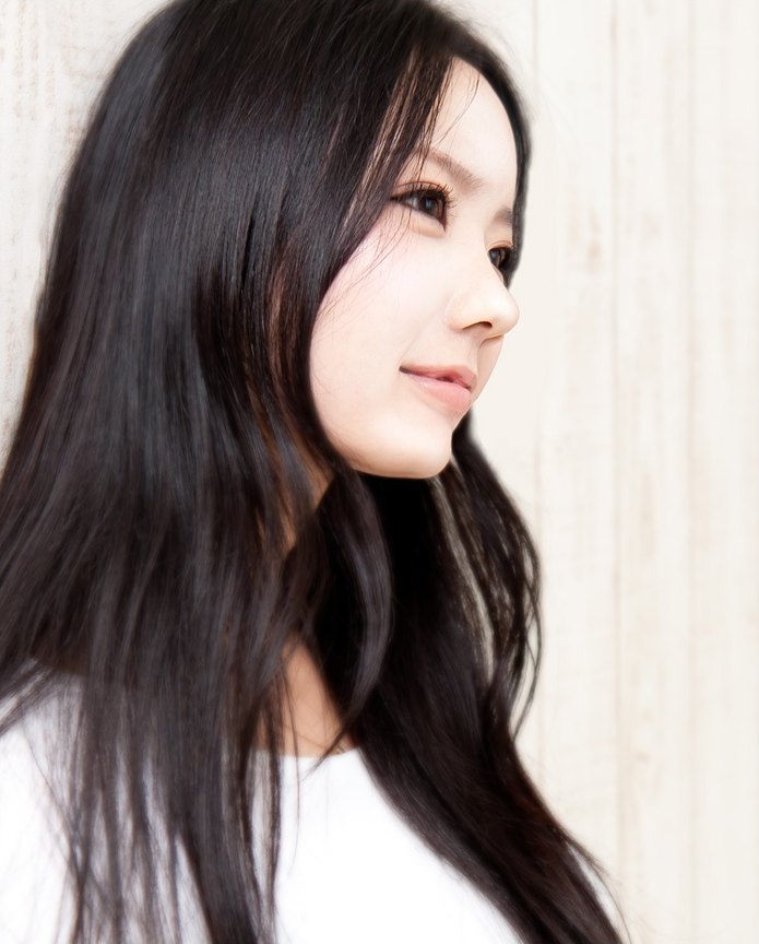 긴 흑발의 여성이 밝은 벽을 배경으로 입체적인 얼굴 옆모습을 드러내며 은은하게 미소 짓는 에이펙스성형외과 귀족수술 클로즈업