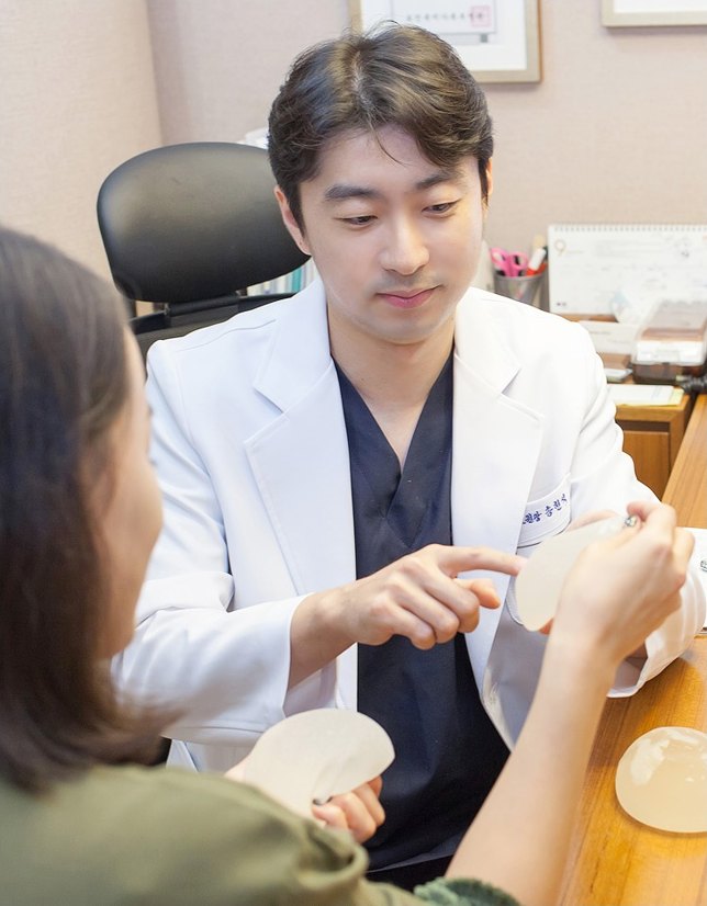 진료실에서 흰 가운을 입은 전문의가 가슴확대 상담 중 환자에게 실리콘 보형물을 보여주며 설명하는 모습