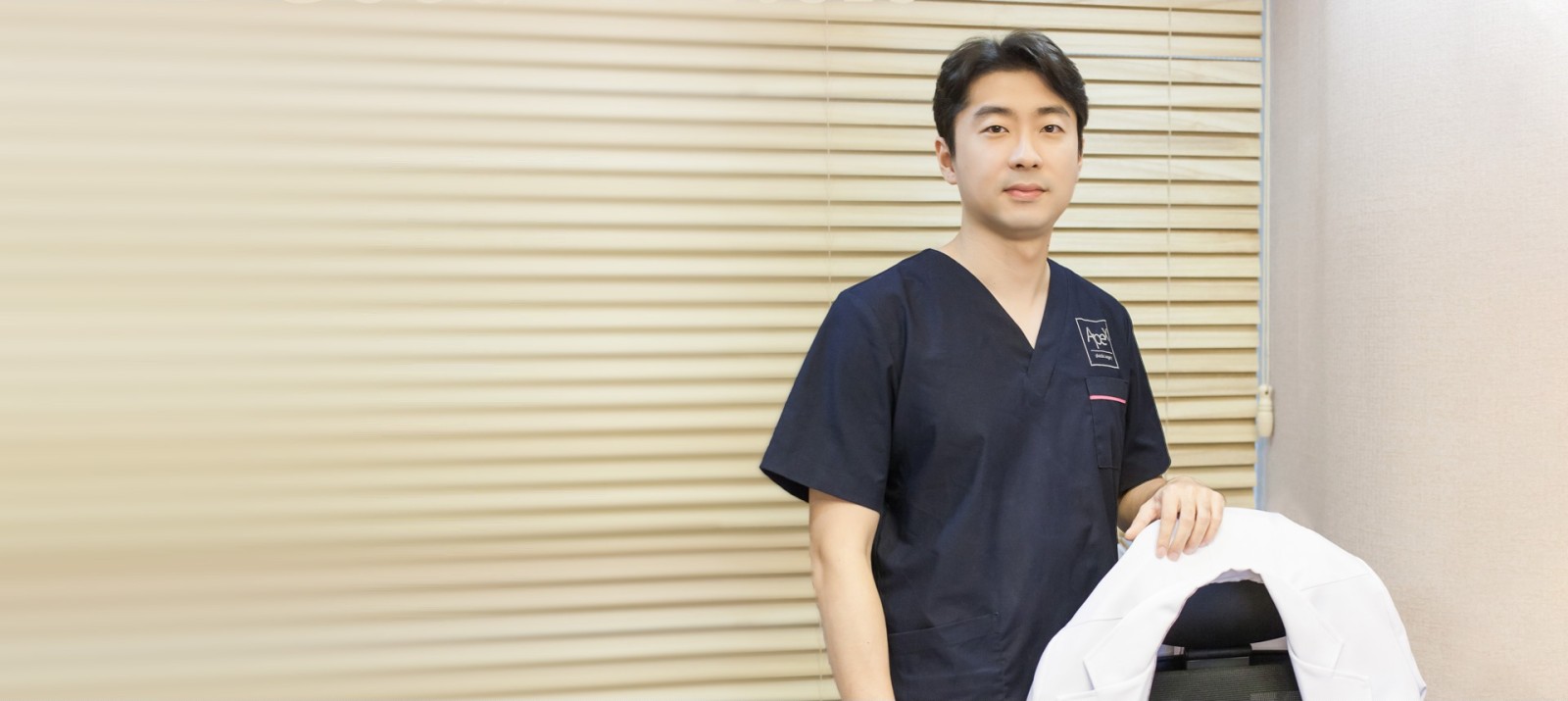 블라인드가 드리운 진료실에서 네이비색 수술복을 입고 의자에 걸친 가운 옆에 서 있는 에이펙스성형외과 의료진의 프로필